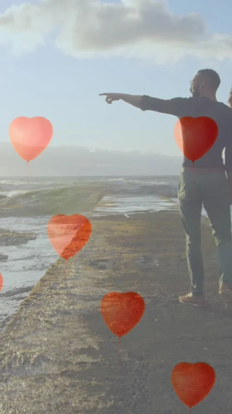 Multiple heart balloons floating against couple pointing towards sea shore Video stock 323781825