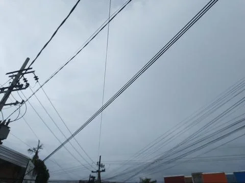 Multiple power lines stretch across a grey cloudy sky with some buildings bel Stock Photos