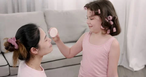 Multiracial girl age 7 using makeup puff to apply powder on asian single mother Stock Footage 314276154