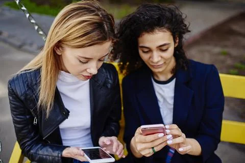 Multiracial ladies sitting on bench while text messaging on mobiles Stock Photos