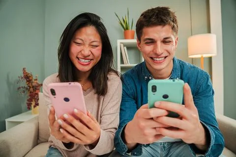 Multiracial young couple using their cellphones sitting on a couch at home Stock Photos