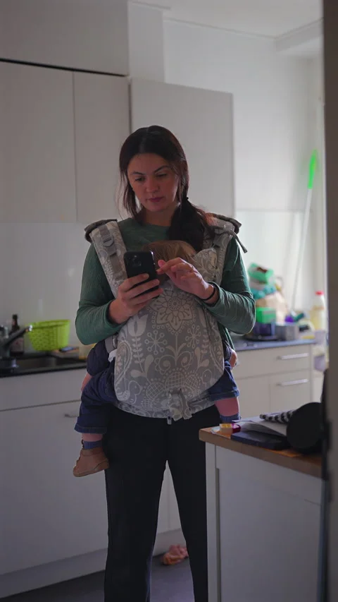 Multitasking mother checks her smartphone while carrying a toddler in a fro.. Video stock 343649447