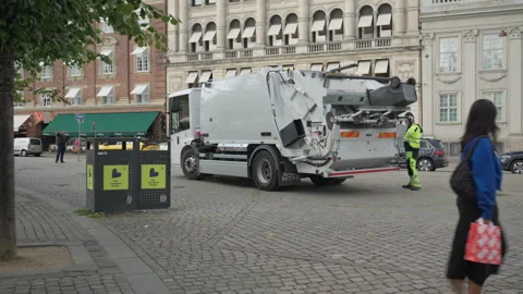 Municipal worker loads a trash bin into a garbage truck on a city square Stock Footage 345590585