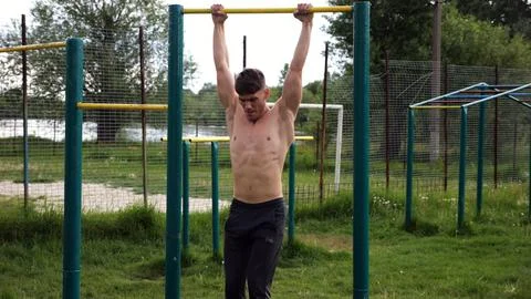 Muscular athlete doing pull ups on horizontal bar at sports ground. Young shi Stock Photos