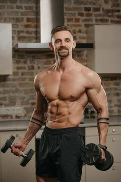 A muscular man with a beard is posing with dumbbells in his apartment. Stock Photos