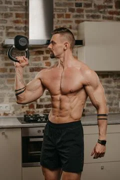 Muscular man with beard is training biceps with a black weight in his apartme Stock Photos