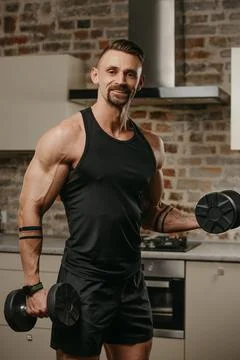 A muscular man with a beard is training biceps with dumbbells in his apartmen Stock Photos