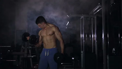 Muscular man doing biceps exercises with dumbbells in the gym. Smoke at the back Video stock 89100277