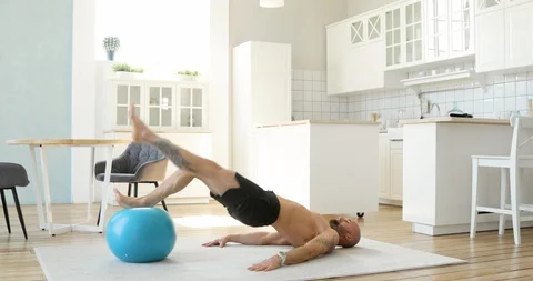 Muscular man doing exercises for buttocks with fitball in living room at home. Stock Footage 129905499