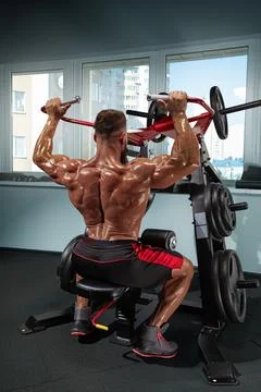 Muscular man performs shoulder press on weight training machine in modern gym Stock Photos