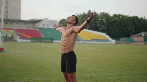 A muscular man stands under drops of water from a sprinkler on a football field Stock-Footage 162379552