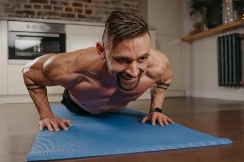 A muscular man with tattoos is doing pushups on a blue yoga mat in his apartm Stock Photos