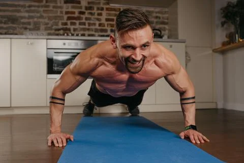 A muscular man with tattoos is doing pushups on a blue yoga mat in his apartm Stock Photos