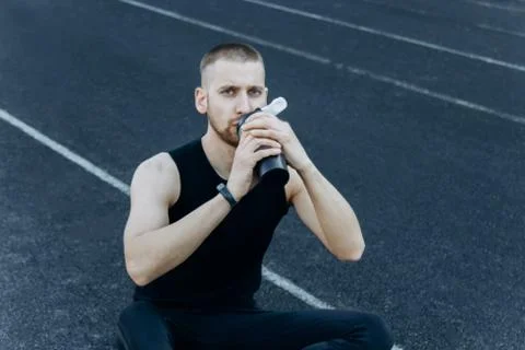 Muscular slender man in training at the stadium drinks water from a sports sh Stock Photos