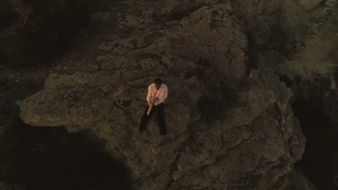 Musician playing saxophone sitting on the edge of a cliff on an island in front Stock Footage