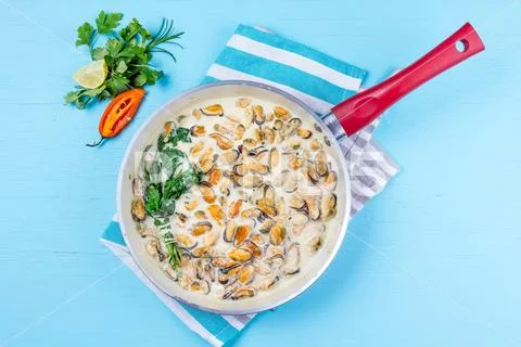 Mussels in milk cream y sauce with aromatic herbs and lemon in red cooking pan Stock Photos
