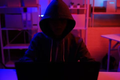 A mysterious hooded hacker working on a computer in a dark room. Stock Photos