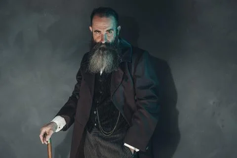 Mysterious vintage beard man in 1900 style fashion with cane against grey wal Stock Photos