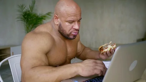 Naked strong man is eating beefburger, sitting at table with notebook and typing Vídeos de archivo 91675343