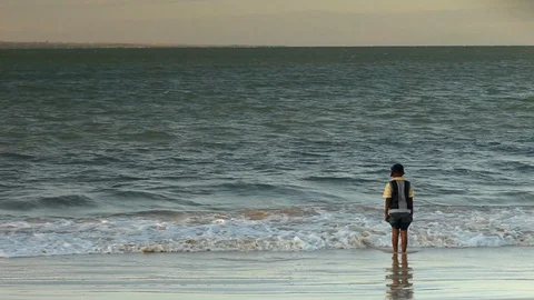 Native boy standing on the beach and enjoying the waves at sunset Video stock 95796791