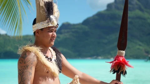 A native islander man blows a conch shell holding a ceremonial spear at a tropic Stock Footage 102579513
