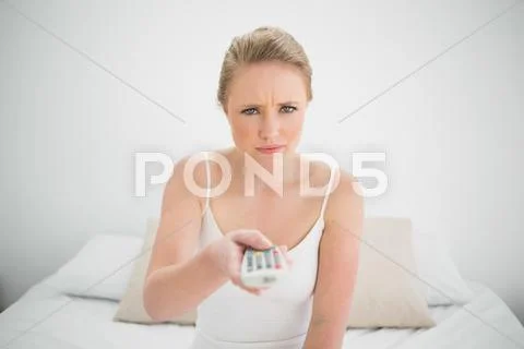 Natural frowning blonde holding remote while sitting on bed Stock Photos