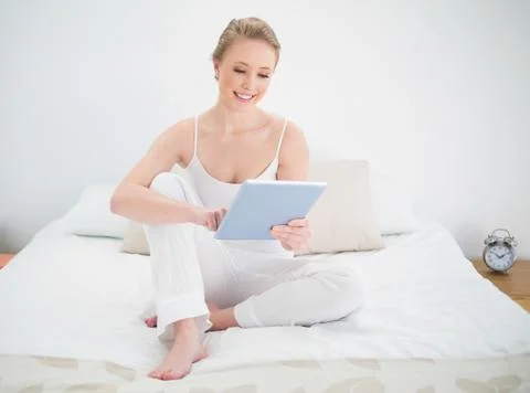 Natural smiling blonde using tablet while sitting on bed Stock Photos