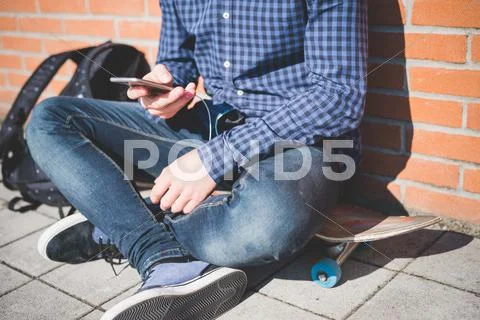 Neck Down View Of Young Male Urban Skateboarder Sitting On Sidewalk Reading