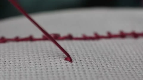  needle piercing the fabric while cross-stitching with red thread.  Stock Footage 231350978