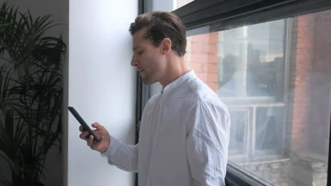 Nervous young man texts on mobile phone and wins with joy near bright window Stock Footage 154121051