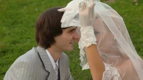 Newly-married couple sits on grass together and kisses under veil of bride Stock Footage