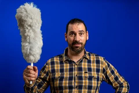 Nice bearded man posing funny with a cleaning duster on blue studio backgroun Stock Photos