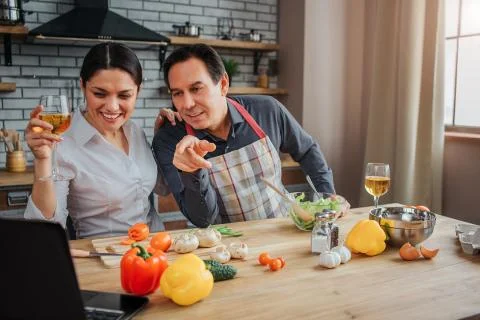 Nice couple sit at table in kitchen and watch on laptop. They hold glasses of Stock Photos
