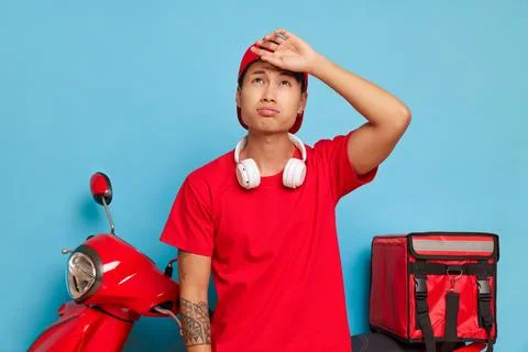 Nice guy in red clothes poses on red scooter with delivery box on blue backdrop Stock Photos