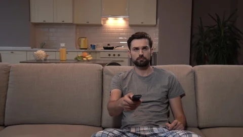 Nice guy sits on sofa and pushes buttons on remote control. He is doing that Stock-Footage 99739160