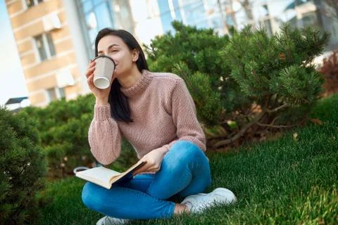 Nice-looking lady drinking coffee while sitting outdoors on a grass with pape Stock Photos