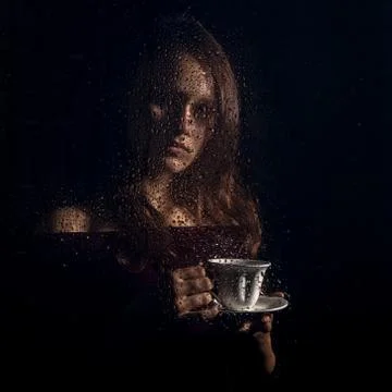 Nice young girl drinks coffee behind the wet glass, raindrops on glass Stock Photos