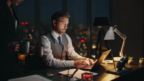 Night employee browsing computer at desk. Smiling woman partner bring coffee Stock Photos