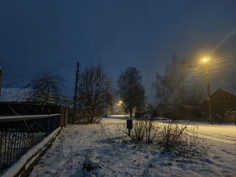 Night falling over a snow-covered rural street as warm lamplight from Stock Photos