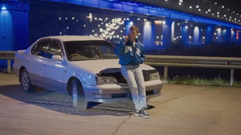Night roadside car under blue bridge, driver leans on hood using phone, waits Stock Footage 324952586