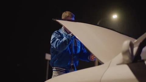 Night roadside white driver inspects hood under streetlight, blue varsity jacket Stock Footage 324954445