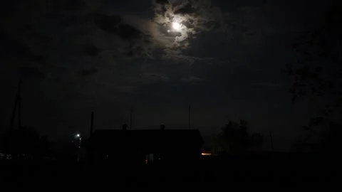 Night time-lapse clouds run against background of moon, on horizon rural house Vidéo 194444324