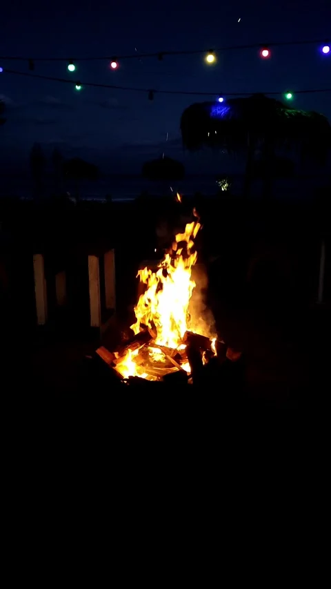 A nighttime beach scene with a bonfire surrounded by ambient colorful string Stock Footage 298040544