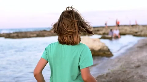 A nine-year-old boy with long, light hair strolls along the seaside, inviting Stock Footage 250817176