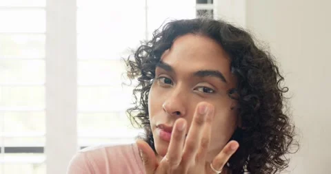 Non-binary adult looking at camera and blending cream by white shutters for Stock Footage 339557369
