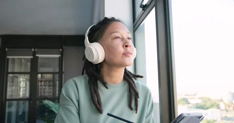 Non-binary African American using stylus on tablet at window, with headphones, Stock Footage 342068489