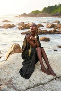 Non-binary black person in luxury dress sits on rocks in ocean. Trans ethnic Stock Photos