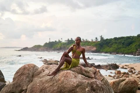 Non-binary black person poses gracefully sitting on top of rock in ocean at Stock Photos