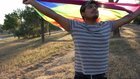 Non-binary guy waving rainbow flag with joy and spin backlighted in full sun 스톡 동영상 250468396
