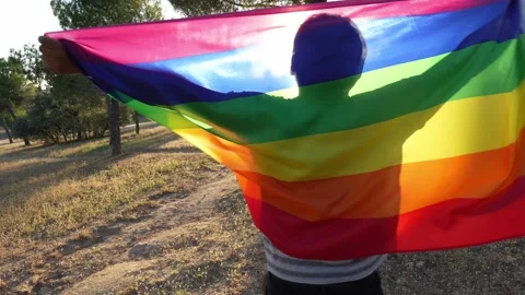 Non-binary guy waving rainbow flag with joy and spin backlighted in full sun 스톡 동영상 250468596